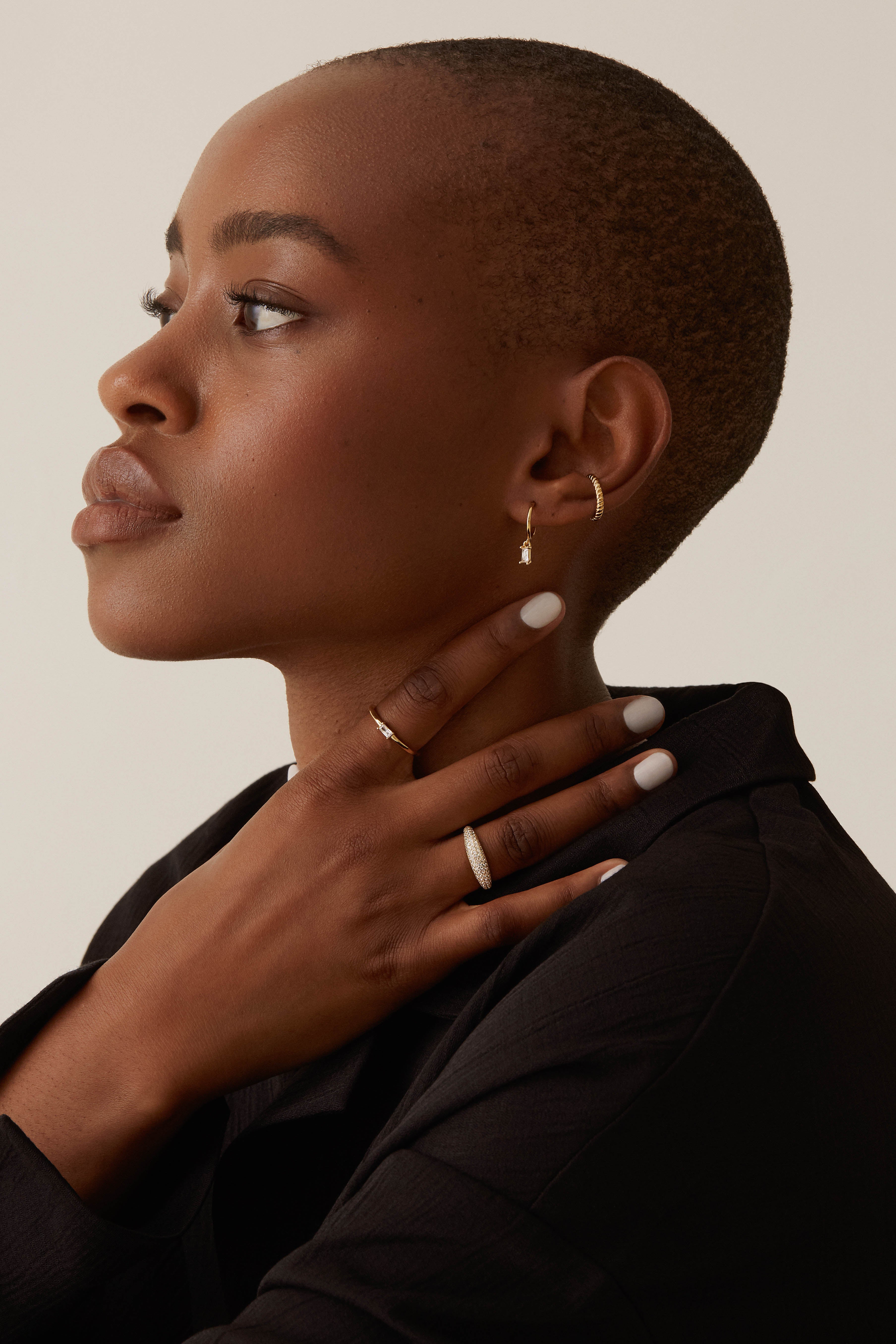 A person with a shaved head and wearing the Alaska Ring in Gold Vermeil by Twenty Compass glances to the side, hand touching their neck. Theyre in a black outfit, showcasing neatly manicured white nails that glisten like cubic zirconia.