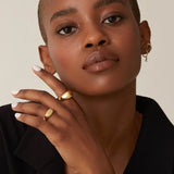 A person with a shaved head dons a black blazer, featuring gold rings and Alaska Earrings in Gold Vermeil by Twenty Compass. Their neutral expression and the cubic zirconia pendants sparkle complete the minimalist look as they pose with one hand near their face against a neutral background.