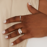A close-up of a hand, adorned in light-colored nail polish and three rings, rests on beige fabric. The central piece is the Twenty Compass Alaska Ring - Silver, featuring a delicate sparkly silver band encrusted with cubic zirconia gems.