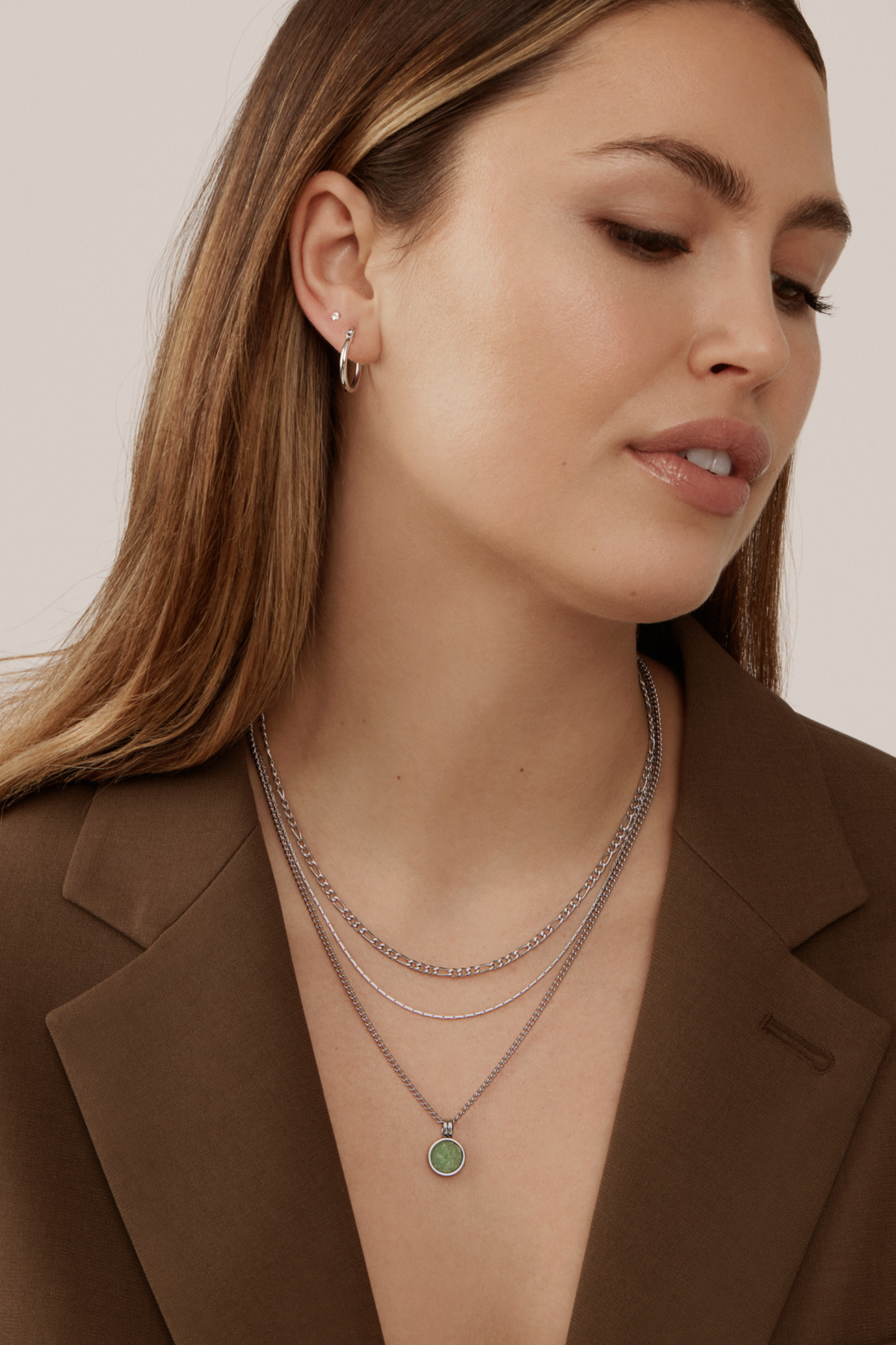 A person with long hair wears a brown blazer, hoop earrings, and layered Andy Necklaces - Silver by Twenty Compass, featuring a green aventurine stone pendant. With a neutral expression, they gaze to the side against a plain background, embodying the essence of the Sarah-Maude Beauchesne collaboration.