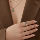 A person in a brown blazer wears a Twenty Compass Andy Necklace - Silver with a green aventurine stone, accessorized by a silver star-designed ring and neatly manicured soft pink nails.