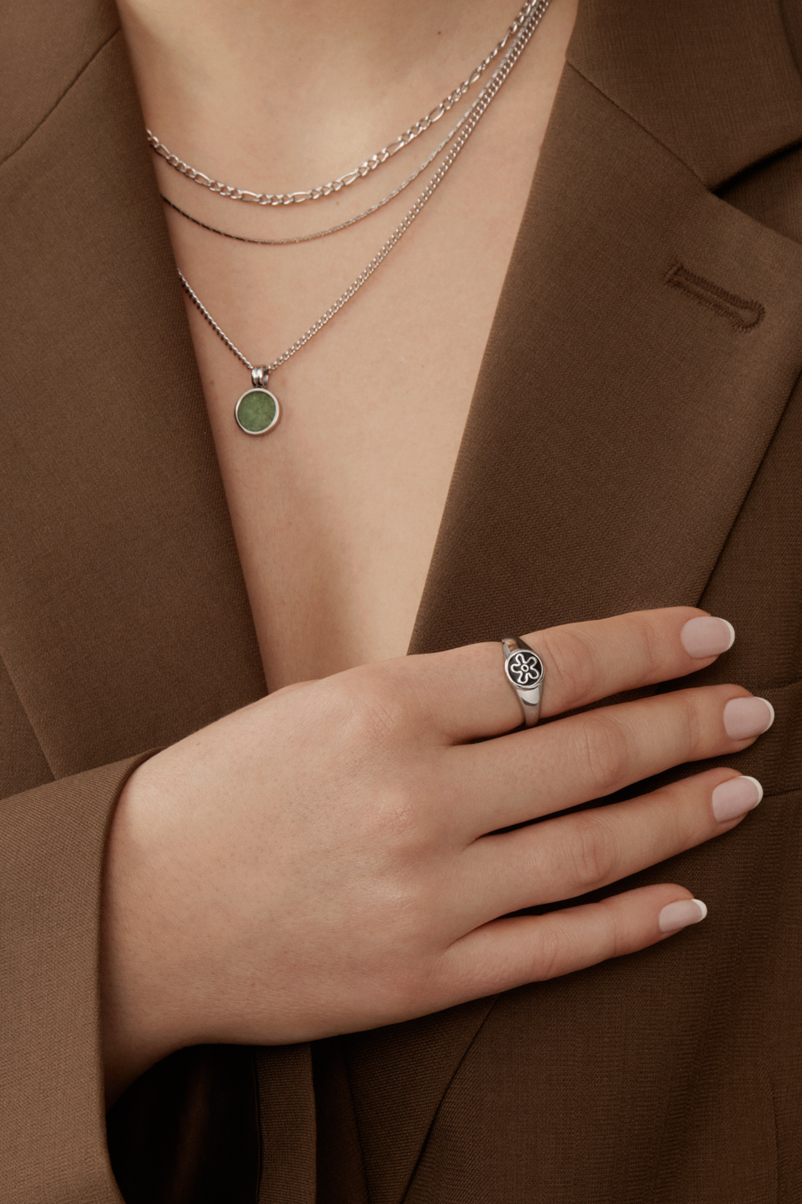 A person in a brown blazer wears a Twenty Compass Andy Necklace - Silver with a green aventurine stone, accessorized by a silver star-designed ring and neatly manicured soft pink nails.