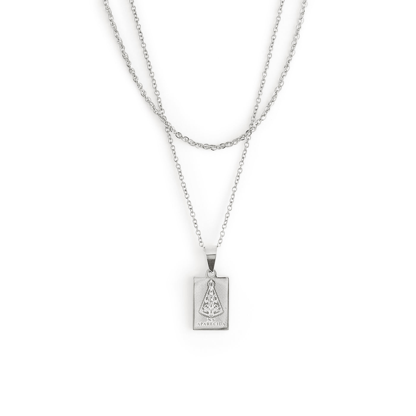 The Aparecida Necklace - Silver by TwentyCompass features a silver double-chain with a rectangular pendant engraved with the Virgin Mary in robes and the text APARECIDA on a white background, serving as a lucky charm. Collier Aparecida - Argent